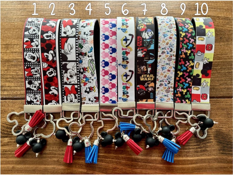 May include: Ten keychains with various designs featuring cartoon characters and Star Wars themes. The keychains have different colored wrist straps with images of Mickey Mouse, Minnie Mouse, and Star Wars characters. Each keychain has a metal clasp and a tassel.