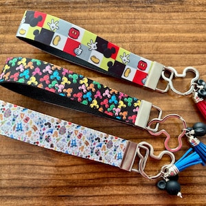 May include: Three wrist keychains with Mickey Mouse-themed designs. One features a black, red, and yellow pattern, another has a black background with colorful Mickey heads, and the third has a white background with cartoon characters. Each has a metal Mickey Mouse-shaped clasp.