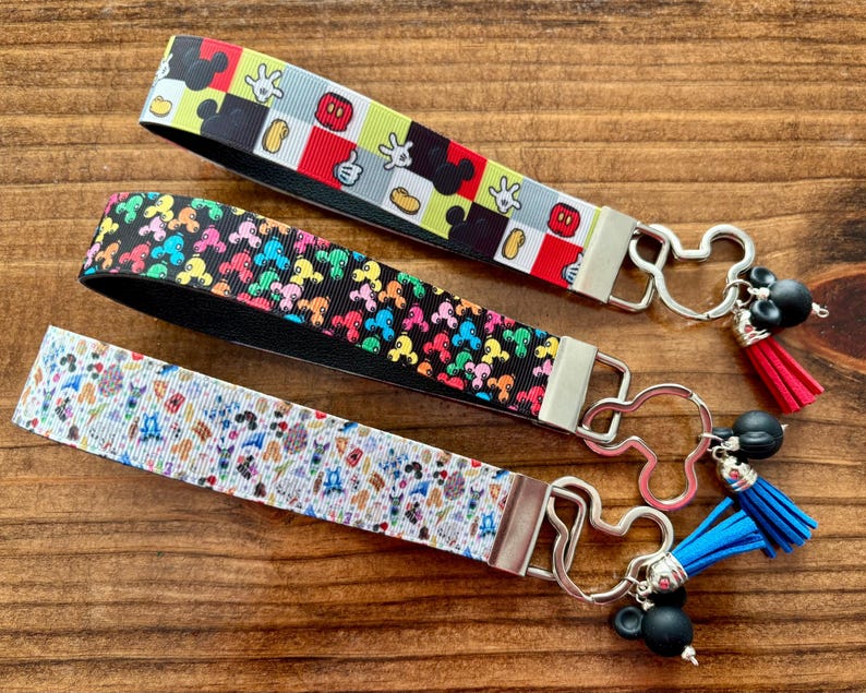 May include: Three wrist keychains with Mickey Mouse-themed designs. One features a black, red, and yellow pattern, another has a black background with colorful Mickey heads, and the third has a white background with cartoon characters. Each has a metal Mickey Mouse-shaped clasp.