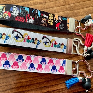 May include: Three keychains with different designs. The top one is black with Star Wars characters. The middle one is white with a Disney design. The bottom one is white with a pink and blue bow pattern. Each keychain has a metal clasp and a tassel.