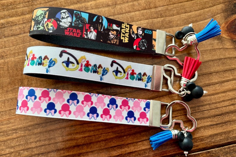 May include: Three keychains with different designs. The top one is black with Star Wars characters. The middle one is white with a Disney design. The bottom one is white with a pink and blue bow pattern. Each keychain has a metal clasp and a tassel.