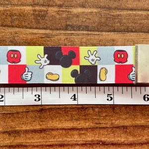 May include: A colorful Mickey Mouse-themed keychain wristlet. The patterned strap features Mickey's gloves, face, and silhouette. The wristlet includes a silver clasp, a black Mickey head charm, and a red tassel. The strap measures approximately 6 inches.