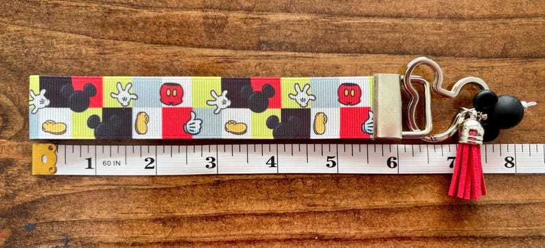 May include: A colorful Mickey Mouse-themed keychain wristlet. The patterned strap features Mickey's gloves, face, and silhouette. The wristlet includes a silver clasp, a black Mickey head charm, and a red tassel. The strap measures approximately 6 inches.