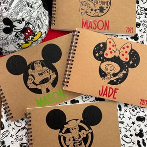 Peut inclure: Trois carnets à spirales marrons avec des oreilles de Mickey Mouse noires et différents motifs de personnages Disney. Les carnets portent les noms "Mason" et "Jade", ainsi que l'année "2025".