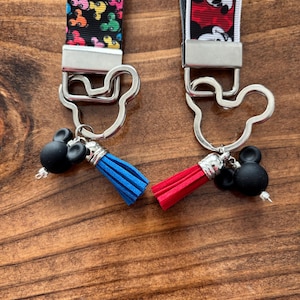 May include: Two keychains with Mickey Mouse-themed designs. One keychain features a black strap with colorful Mickey Mouse heads, a silver clasp, a black Mickey Mouse head charm, and a blue tassel. The other has a red and white strap with Mickey Mouse, a silver clasp, a black Mickey Mouse head charm, and a red tassel.