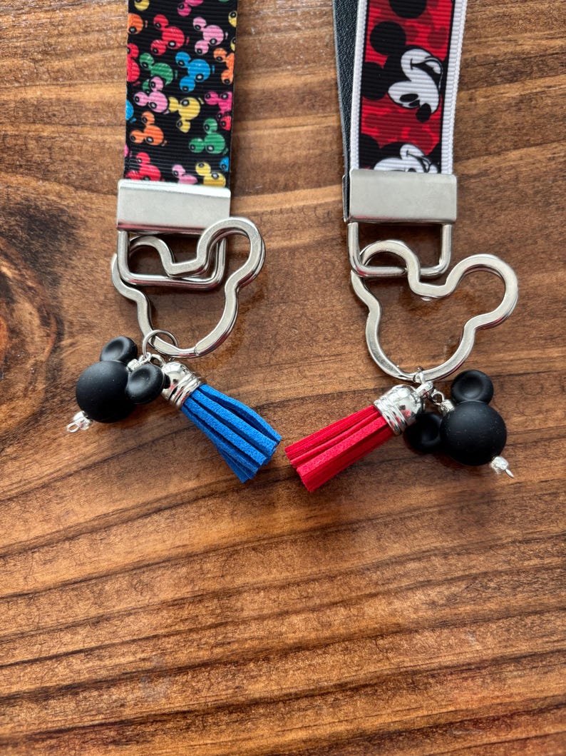 May include: Two keychains with Mickey Mouse-themed designs. One keychain features a black strap with colorful Mickey Mouse heads, a silver clasp, a black Mickey Mouse head charm, and a blue tassel. The other has a red and white strap with Mickey Mouse, a silver clasp, a black Mickey Mouse head charm, and a red tassel.