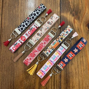 May include: A collection of key fobs with various designs, including cow print, Diet Coke branding, coffee cups, floral patterns, cartoon characters, and a Houston Astros logo. Each fob has a metal clasp and a tassel.