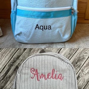 May include: Two seersucker backpacks. One is light blue with the word "Aqua" and a white zipper. The other is gray and white striped with the name "Amelia" in pink and the word "Gray".