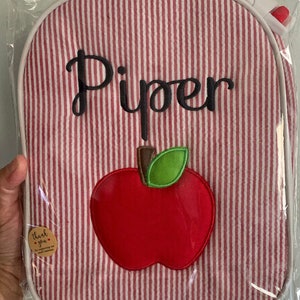 Op de afbeelding: Rode en witte gestreepte lunchtas met een rode appel en een groene bladapplicatie. De tas is gepersonaliseerd met de naam "Piper" in zwarte borduurwerk.