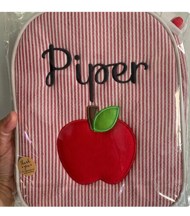 Op de afbeelding: Rode en witte gestreepte lunchtas met een rode appel en een groene bladapplicatie. De tas is gepersonaliseerd met de naam "Piper" in zwarte borduurwerk.