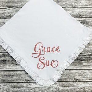 May include: White baby blanket with a ruffled edge and the name "Grace Sue" embroidered in pink.