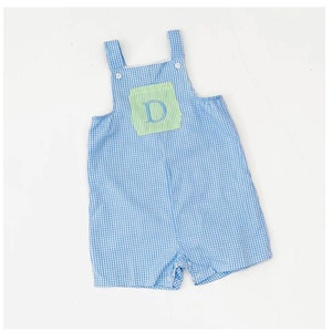 Pode incluir: Macacão de bebé em padrão xadrez vichy azul e branco, com alças ajustáveis e fechos de pressão. Apresenta um bolso verde bordado com a letra "D". Um traje clássico e confortável para um bebé.