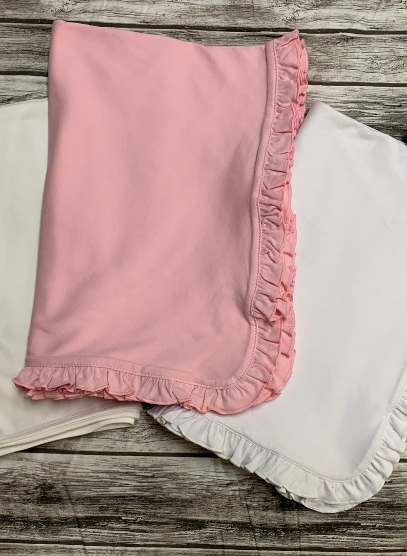 May include: Two baby blankets, one pink and one white, with ruffled edges. The pink blanket is folded on top of the white blanket.