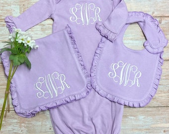 May include: A set of three lavender baby items with white embroidered initials "SWR". The set includes a long-sleeved gown, a burp cloth, and a bib, all with ruffled edges.