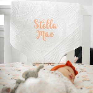 May include: A white quilted baby blanket with the name "Stella Mae" embroidered in peach on it. A baby is sleeping on a floral crib sheet.