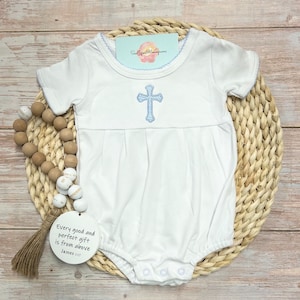 Pode incluir: Body branco para bebé com um bordado de cruz azul claro na frente. O body tem mangas curtas e acabamentos em azul claro. Uma etiqueta com o texto "Every good and perfect gift is from above James 1:17" está anexada.