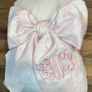 Könnte beinhalten: Eine weiße Decke mit einer großen rosa Schleife mit Toile-de-Jouy-Muster. Die Schleife ist mit einem rosa Monogramm bestickt. Die Decke ist gefaltet und mit der Schleife gebunden, was eine dekorative Präsentation ergibt.