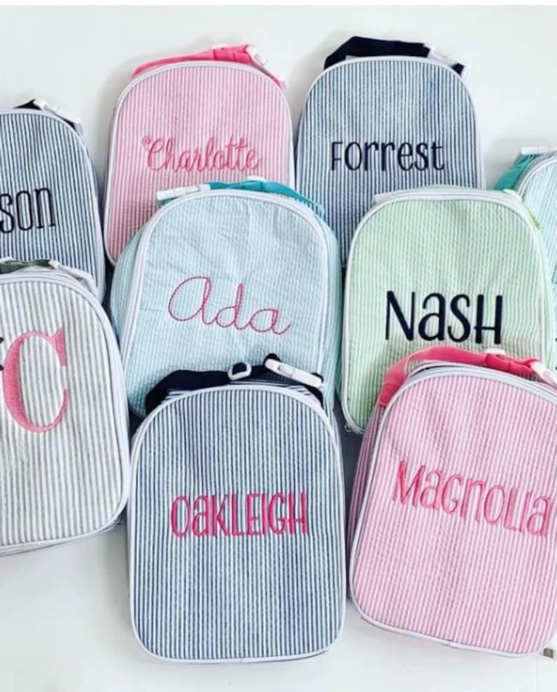 Op de afbeelding: Een verzameling van zeven gepersonaliseerde lunchtassen in verschillende kleuren en patronen. De tassen zijn gemaakt van gestreept stof en hebben verschillende namen geborduurd, waaronder Charlotte, Forrest, Ada, Nash, Oakleigh en Magnolia.