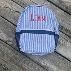 May include: A blue and white striped backpack with the name "LIAM" embroidered in red. The backpack has a navy blue trim and mesh side pockets. The bag is made of a seersucker fabric.