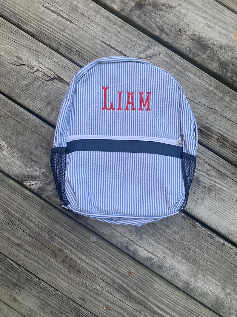 May include: A blue and white striped backpack with the name "LIAM" embroidered in red. The backpack has a navy blue trim and mesh side pockets. The bag is made of a seersucker fabric.