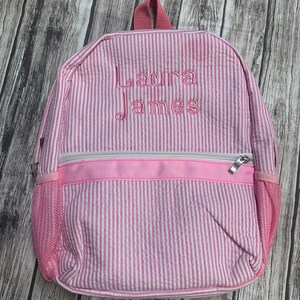 May include: Pink and white striped backpack with the name "Laura James" embroidered in pink thread. The backpack has a front zippered pocket, mesh side pocket, and a top handle. The background is a wooden surface.