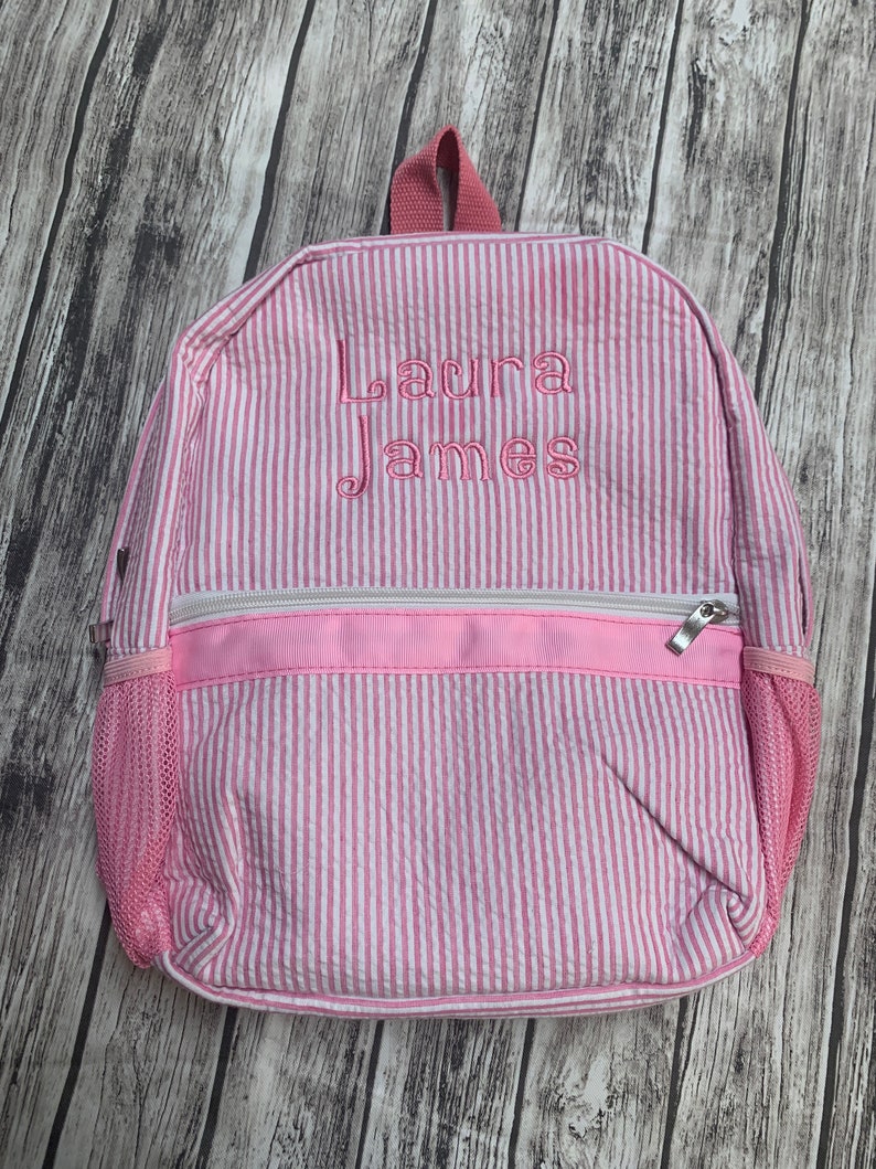 May include: Pink and white striped backpack with the name "Laura James" embroidered in pink thread. The backpack has a front zippered pocket, mesh side pocket, and a top handle. The background is a wooden surface.
