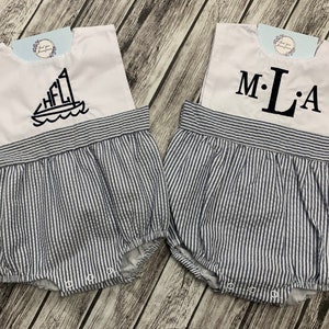 May include: Two baby rompers. Each has a white top with a blue and black sailboat or initials design. The bottom is blue and white striped. The rompers have snap closures.