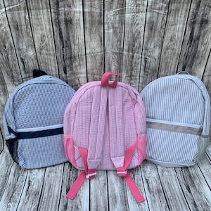 May include: Three seersucker backpacks in blue, pink, and gray, each with vertical stripes. The blue backpack has a navy blue accent, the pink has pink straps, and the gray has a gray accent. All have adjustable straps and side pockets.
