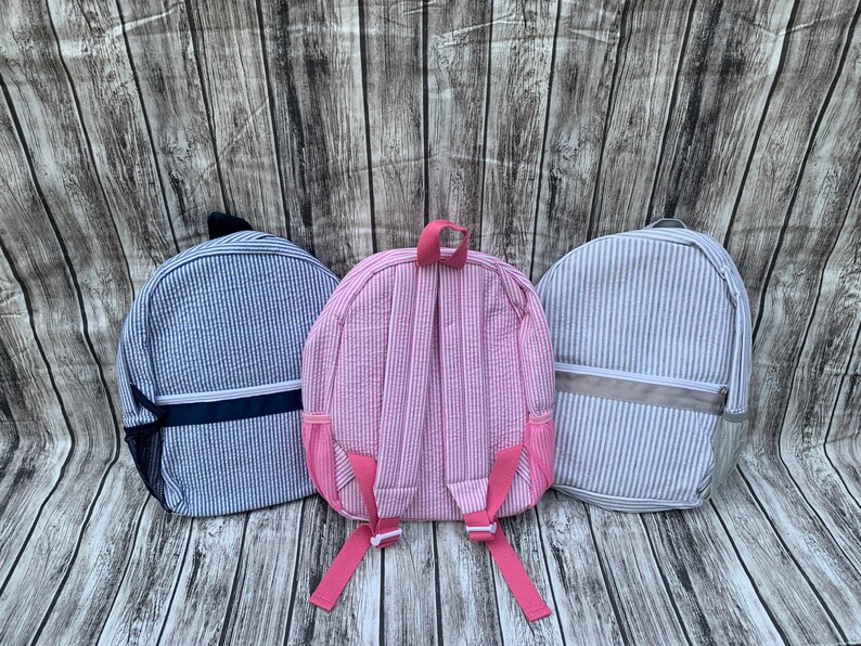 May include: Three seersucker backpacks in blue, pink, and gray, each with vertical stripes. The blue backpack has a navy blue accent, the pink has pink straps, and the gray has a gray accent. All have adjustable straps and side pockets.