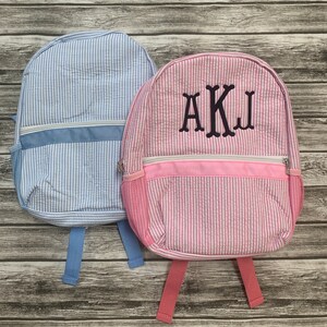 May include: Two seersucker backpacks. One is light blue with a white zipper and trim, and the other is pink with a white zipper and trim. The pink backpack has the embroidered letters AKJ. Both have side mesh pockets and adjustable straps.