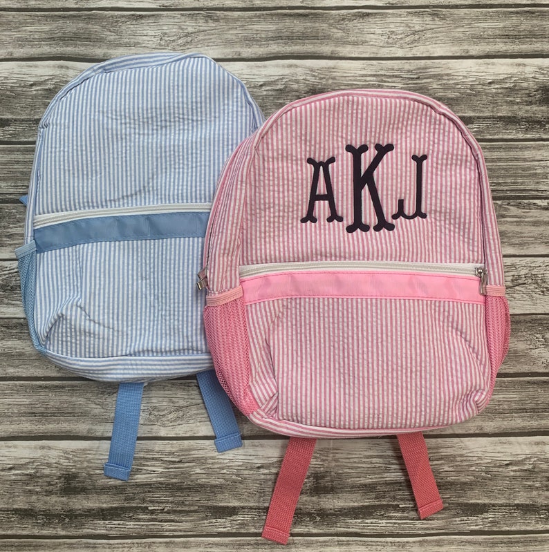 May include: Two seersucker backpacks. One is light blue with a white zipper and trim, and the other is pink with a white zipper and trim. The pink backpack has the embroidered letters AKJ. Both have side mesh pockets and adjustable straps.