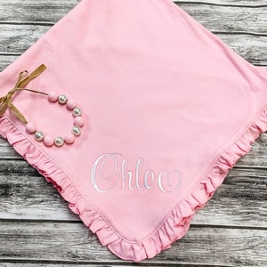 May include: A pink baby blanket with a ruffled edge and the name "Chloe" embroidered in white. A white beaded necklace is on top of the blanket.