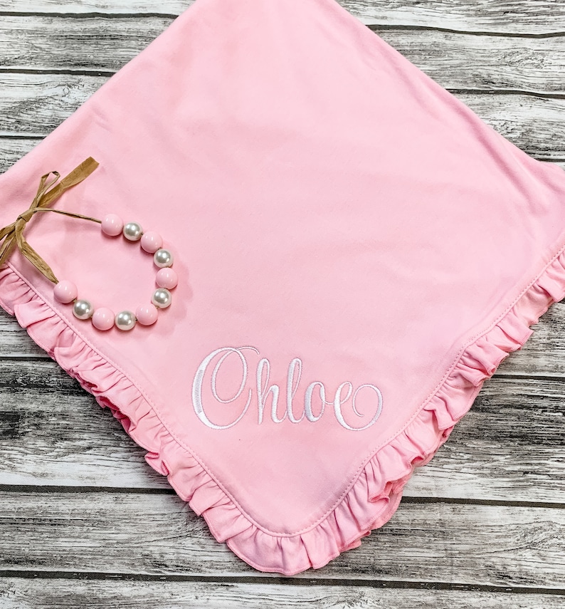 May include: A pink baby blanket with a ruffled edge and the name "Chloe" embroidered in white. A white beaded necklace is on top of the blanket.