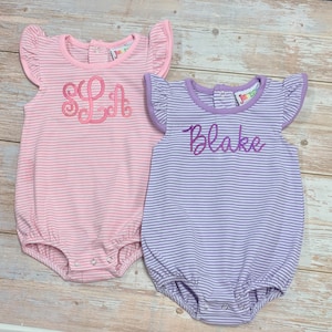 Pode incluir: Dois macacões de bebé. Um é rosa com um monograma "SL" em fio rosa, o outro é lavanda com o nome "Blake" bordado em fio roxo. Ambos têm padrões listrados, mangas com babados e fechos de pressão.