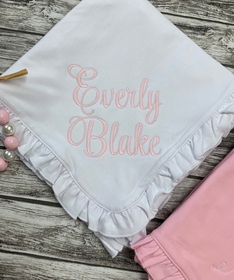 May include: White baby blanket with a pink ruffled edge and the name "Everly Blake" embroidered in pink script.