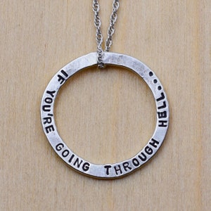 May include: A silver pendant necklace with a circular design. The pendant has the stamped text "IF YOU'RE GOING THROUGH HELL". The necklace hangs on a delicate silver chain, set against a light wood grain background.