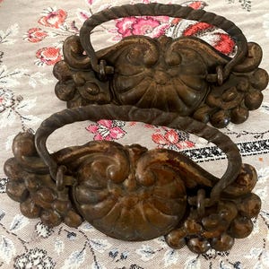 May include: Two antique bronze drawer pulls with ornate floral and scrollwork designs. Each pull features a curved handle and decorative base with a central shell-like motif and clusters of small, round details. The pulls are set against a floral patterned background.