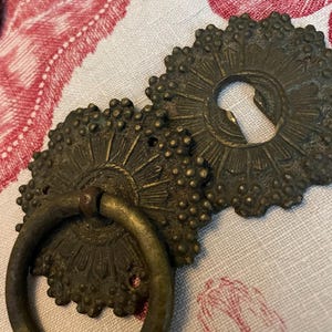 May include: Two antique brass furniture hardware pieces featuring a dark, aged patina. One piece is a circular escutcheon plate with a keyhole, and the other is a matching circular backplate with a hinged, round pull ring attached to the center.
