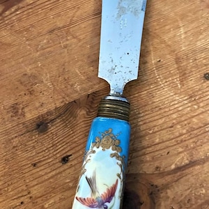 以下が含まれることがあります： ステンレス鋼の刃と青い磁器のハンドルが付いた、ヴィンテージのバターナイフ。 ハンドルには、金色のアクセントが付いた鳥の絵柄が描かれており、この道具にエレガントさを加えています。