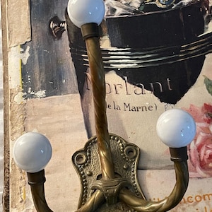 May include: A triple-pronged wall hook featuring a textured brass base and three curved, twisted brass arms. Each arm terminates in a smooth, round white ceramic knob. The hook is designed for mounting with visible screw holes at the top and bottom.