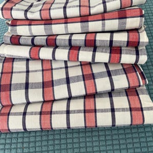 Puede incluir: Una pila de servilletas de tela blanca dobladas con un patrón de cuadros rojos y azul marino. Las servilletas están cuidadosamente dispuestas, mostrando el diseño de cuadrícula repetitivo. La tela parece ser de algodón o lino.