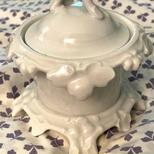 May include: A white ceramic sugar bowl with a matching lid featuring a branch-like handle. The bowl and lid are decorated with embossed leaf motifs along the rim and base, creating a textured appearance with a glossy finish and smooth surface.