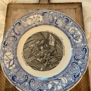 Könnte beinhalten: Ein blauer und weißer Keramikteller mit einer Schwarzweißillustration einer historischen Szene. Der Teller hat einen dekorativen Rand mit floralen und geometrischen Mustern. Der Text auf dem Teller lautet "NUTOIRE DE JEANNE D'ARC" und "CHARLES OF ELLE IS FRA MERA RIMS".
