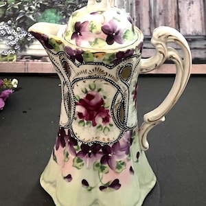 Puede incluir: Cafetera antigua de porcelana con tapa, con un diseño floral en tonos morados, rosas y verdes. La cafetera tiene un asa y un pico curvos, con detalles dorados. El diseño incluye rosas y otras flores.