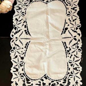 May include: White, rectangular lace table runner with a floral and leaf design. The runner features two oval-shaped solid areas in the center. The edges are intricately cut with a delicate pattern. The runner is placed on a dark surface.