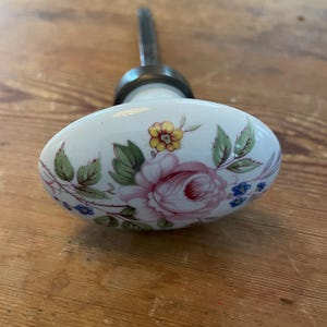 May include: White ceramic door knob with a floral design featuring pink roses, yellow and blue flowers, and green leaves. The knob has a metal stem and a brass collar. The floral pattern is hand-painted on the oval-shaped knob.