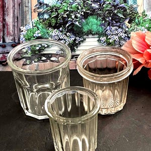 May include: Three clear, vintage glass jars of varying sizes. The jars have a ribbed design and a thick, rounded rim. The background features a floral arrangement and a window.