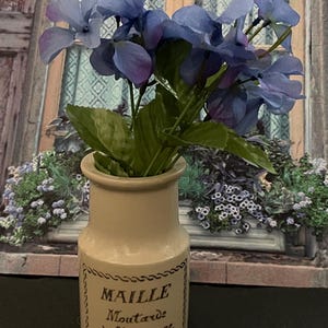 Puede incluir: Un pequeño tarro de cerámica blanquecina con el texto "MAILLE Moutarde à l'ancienne" contiene un ramo de flores azules y moradas con hojas verdes. El tarro es cilíndrico y tiene un borde decorativo.