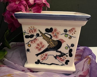 Op de afbeelding: Een witte keramische plantenpot met een blauwe rand en een bloemenontwerp met een vogel in de vlucht. De pot heeft een vierkante vorm en is versierd met roze bloemen en groene bladeren.
