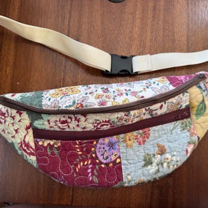 May include: Quilted fanny pack with a patchwork design featuring floral patterns in various colors. The bag has a cream-colored adjustable strap with a black buckle and a brown zipper.